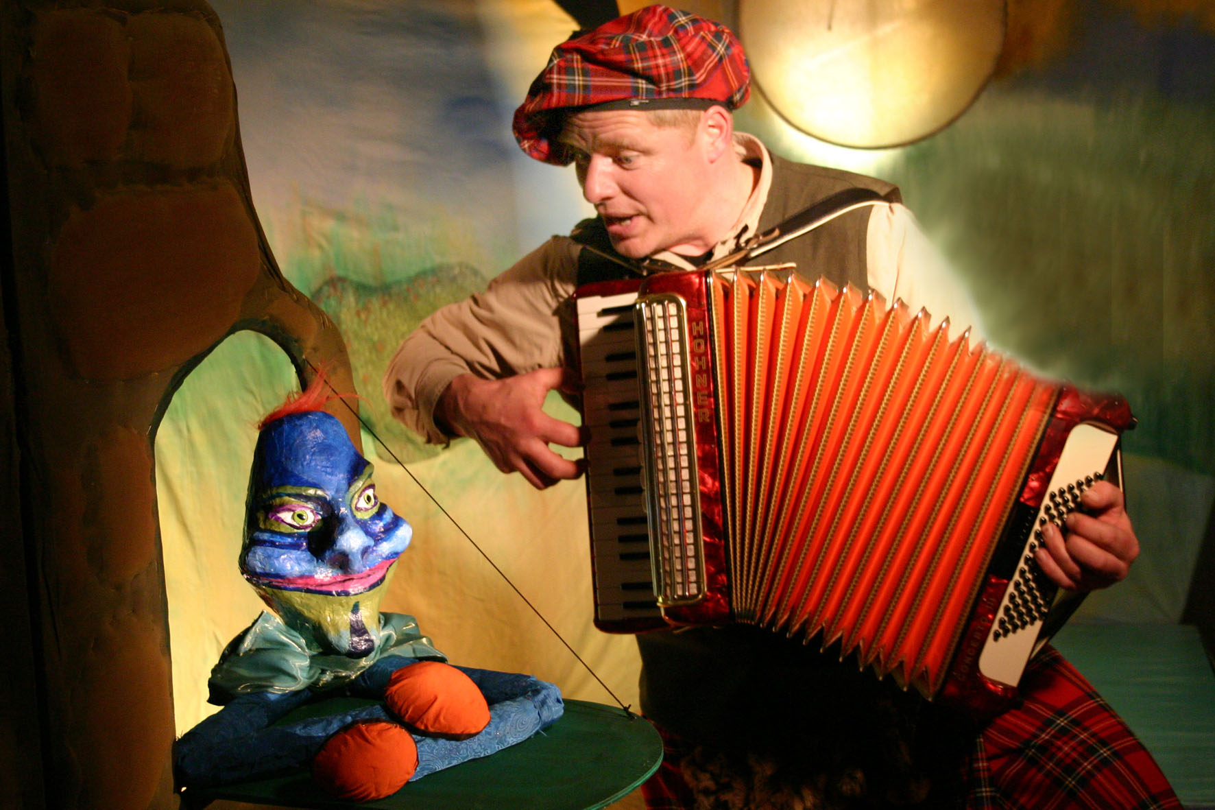 Ein Mann mit bunter Kappe und Akkordeon sitzt neben einem Stofftier.