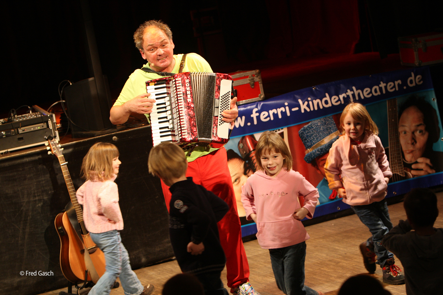 Ein Kinderliedermacher singt und spielt Akkordeon. Um ihn herum tanzen Kinder